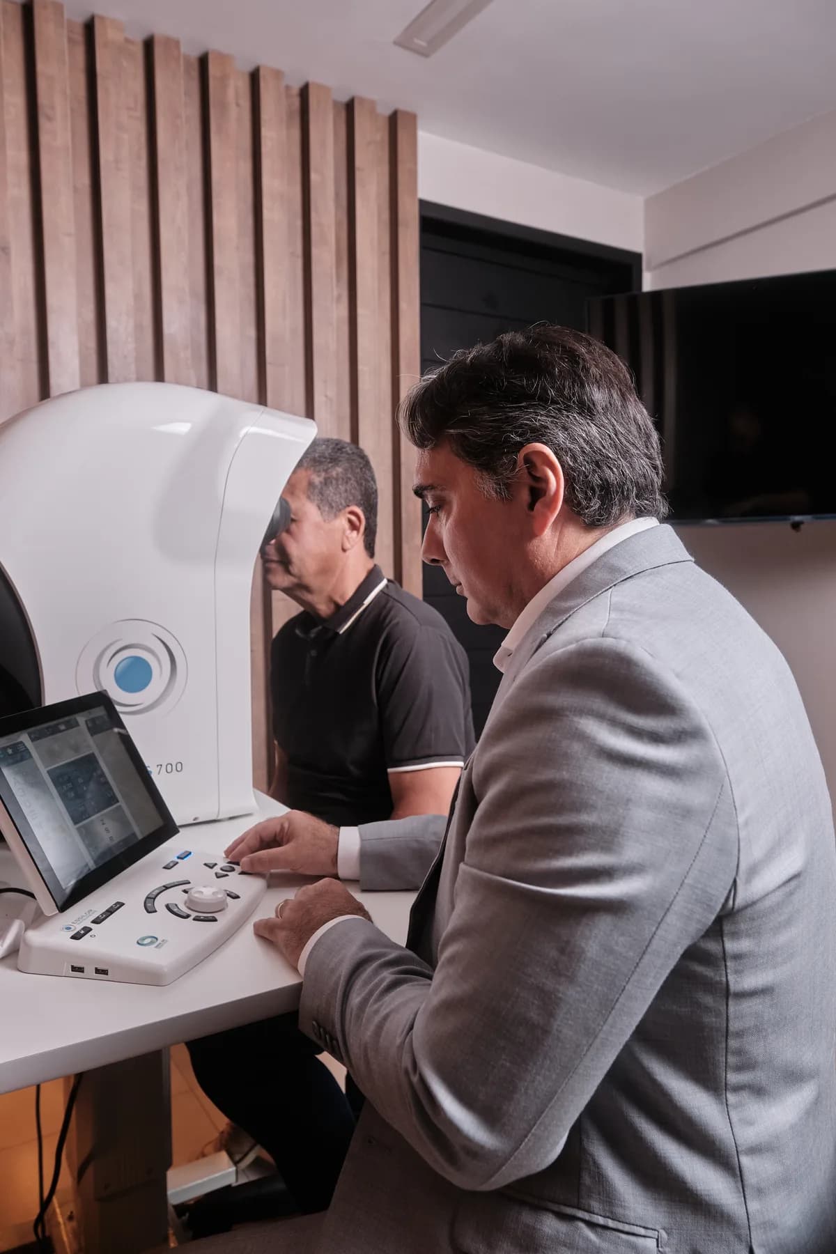 Dr. João Marcelo Lyra conduzindo exame de um paciente no equipamento de diagnóstico, em sala com painel de madeira